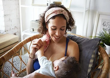 Mulher amamentando bebê. Ela segura a mão dele, eles se olham e ela sorri.